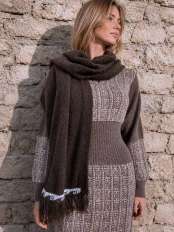 Mujer rubia ante un muro de piedra, con vestido de punto y chal bufanda marrón de lana de alpaca con flecos, modelo Kora.